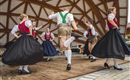 Eine Gruppe von Tänzern in traditioneller bayerischer Tracht tanzt auf einer Bühne. Die Frauen tragen lange schwarze Röcke und die Männer kurze Hosen mit Socken.