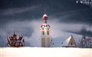 Ein Kirchturm mit einer roten Kuppel ragt aus einer schneebedeckten Landschaft hervor. Im Hintergrund sind verschneite Hügel und Nebel zu sehen.