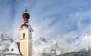 Eine hübsche Kirche mit einer charakteristischen Kuppel steht im Schnee. Im Hintergrund sind majestätische Berge und ein blauer Himmel zu sehen.