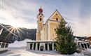 Eine schöne Kirche mit rotem Kirchturm und gelber Fassade im Schnee. Der Sonnenstrahl verleiht der winterlichen Landschaft einen besonderen Glanz.
