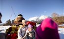 Eine Familie sitzt auf einem Schlitten im Schnee, umgeben von einer winterlichen Landschaft. Im Hintergrund sind Berge unter einem klaren blauen Himmel zu sehen.