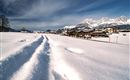 Eine schneebedeckte Landschaft mit klaren Fußspuren im Schnee. Im Hintergrund sind Berge und ein schöner Ort zu sehen.