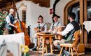 Eine Gruppe von Musikern spielt traditionelle Instrumente in einem gemütlichen Außenbereich. Die Musiker tragen bayerische Trachten und umgeben sich mit schöner Dekoration.