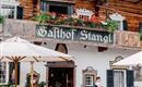 Ein traditionelles Gasthaus mit holzverkleideter Fassade und blühenden Pflanzen. Tische im Freien unter Sonnenschirmen bieten einen einladenden Platz zum Verweilen.