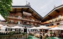 Ein traditionelles alpenländisches Gasthaus mit Holzfassade und blühenden Balkonen. Gäste sitzen unter Sonnenschirmen im Freien und genießen die Atmosphäre.