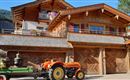 Ein traditionelles Holzhaus mit einem Balkon und Blumen. Vor dem Haus steht ein oranger Traktor und ein Wagen mit Blumen.