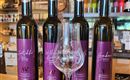 Vier Flaschen mit Wein, die nebeneinander stehen, und ein Weinglas im Vordergrund. Die Etiketten sind in lila gehalten und enthalten verschiedene Namen.