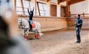 Eine Reiterin zeigt eine Akrobatikübung auf einem weißen Pferd in einer Reitarena. Der Trainer steht daneben und beobachtet die Übung.