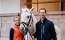 Eine Frau und ein Mann stehen mit einem weißen Pferd in einem Reitstall. Der Mann trägt eine dunkle Jacke und die Frau eine orange Weste.