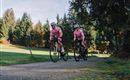 Zwei Radfahrer in rosa Trikots fahren auf einem Schotterweg. Im Hintergrund sind grüne Wiesen und Bäume zu sehen.