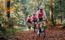 Drei Radfahrer in pinken Trikots fahren auf einem Waldweg. Der Boden ist mit buntem Laub bedeckt und die Bäume umgeben sie.