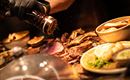 Ein köstliches Stück Fleisch wird frisch gewürzt. Dazu liegen Beilagen wie Kartoffeln und Gemüse bereit.