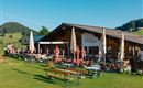 Ein gemütliches Restaurant in der Alpenlandschaft mit einer großen Terrasse. Umgeben von grünen Wiesen und bewaldeten Hügeln.