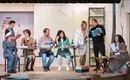 A theater performance with several actors on stage. The performers are in vibrant costumes and interact at a table.
