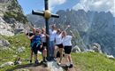 A group of people stands in front of a mountain cross in the mountains. They are smiling and happily posing for the photo.