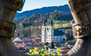 Eine malerische Aussicht auf eine Kirche in einer kleinen Stadt, umgeben von grünen Hügeln. Im Vordergrund ist ein dekorativer Holzrahmen zu sehen.