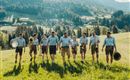 Een groep mannen in Beierse klederdracht marcheert over een groene weide. Op de achtergrond zijn bergen en bossen te zien.
