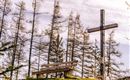 A wooden bench stands next to a wooden cross, surrounded by bare trees. The sky is slightly overcast.
