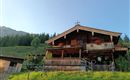 Ein traditionelles Holzhaus in den Bergen, umgeben von grünen Wiesen. Vor dem Balkon sind bunte Blumen und eine Kreuzdekoration zu sehen.