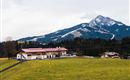 Ein malerisches Landschaftsbild mit einem Bauernhaus im Vordergrund und einem majestätischen Berg im Hintergrund. Der Himmel ist bewölkt und die Wiesen sind grün.