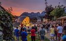 A picturesque village view at night with mountains in the background. People gather in the square under bright lights.