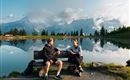 Zwei Personen sitzen auf einer Bank am Ufer eines Sees. Im Hintergrund sind Berge und ein klarer Himmel zu sehen.