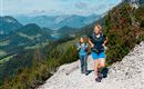 Zwei Frauen wandern auf einem schmalen Pfad in den Bergen. Im Hintergrund erstreckt sich eine malerische Landschaft mit Bergen und Tälern.