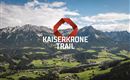 An impressive mountain landscape with green meadows and high mountains in the background. In the foreground stands the logo for the Kaiserkronen-Trail.