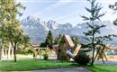 A beautiful playground in a park with a unique play equipment and shade-giving trees. In the background, impressive mountains are visible.