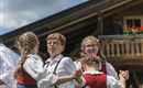 Kinder in traditioneller Tracht tanzen zusammen vor einem rustikalen Holzhaus. Der Himmel ist blau und die Stimmung ist fröhlich.