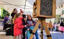 A group of children stands around an old camera and looks curiously. In the background, more people and a festive atmosphere can be seen.