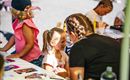 Ein Kind bekommt ein Gesichtsmalerei von einer Betreuerin. Im Hintergrund sind weitere Kinder beim Malen zu sehen.