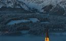 Eine idyllische Berglandschaft mit schneebedeckten Gipfeln und einem kleinen Dorf. Die Kirche im Dorf leuchtet sanft im Dunkeln.