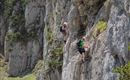 Two climbers rappelling down a rock face. The surroundings are green and mountainous, and the sun is shining.
