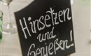 Ein stilvolles Tischsetting mit Gläsern und bunten Kerzen. Ein Schild lädt mit „Hinsetzen und Genießen!“ ein.