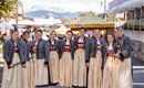 Eine Gruppe von Mädchen und Frauen in traditionellen Trachten steht lächelnd zusammen. Im Hintergrund sind Berge und ein Festzelt zu sehen.