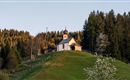 Eine kleine Kirche auf einem Hügel, umgeben von Bäumen. Die Landschaft ist grün und die Atmosphäre ist ruhig.