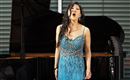 Eine Sängerin in einem blauen, glitzernden Kleid steht vor einem Klavier und singt leidenschaftlich. Im Hintergrund sind Fenster mit horizontalen Jalousien sichtbar.