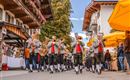 A music band in traditional costumes marches through a festive street. In the background, stands and colorful sun umbrellas can be seen.