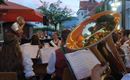 A brass band is playing outdoors in the evening. In the foreground, a musician with a shiny tuba can be seen.