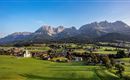 A picturesque landscape with green meadows and a small village. In the background, majestic mountains rise under a clear sky.