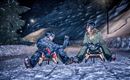 Zwei Personen sitzen im Schnee auf Rodeln und lachen sich an. Die Umgebung ist nachts und mit Schnee bedeckt, umgeben von schneebedeckten Bäumen.
