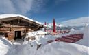 A cozy mountain cabin in the snow with red tables outside. In the background, majestic mountains are visible under a blue sky.
