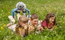 Four children are lying on a meadow and looking at the grass with a magnifying glass. A colorful, creatively designed creature is observing them from behind.