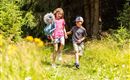 Two children are happily running on a woodland path, surrounded by green plants and flowers. The girl is carrying a toy and smiling as she plays with the boy.