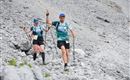 Two athletes are walking with hiking poles over rocky terrain. They are wearing light sports clothing and appear motivated.