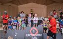 A group of children stands on a stage at the Kaiserkrone Trail. They hold medals and trophies while adults cheer them on.