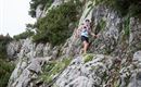 A man is climbing on a rocky path in the mountains. The surroundings are forested and cloudy.