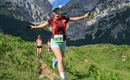 A cheerful runner wearing sunglasses and a backpack enjoys the mountain landscape. In the background, majestic mountains and a clear sky can be seen.