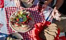 Een kleurrijke plaat met verschillende vlees- en kaaswaren, verse groenten en brood. Op de achtergrond zijn drankjes en andere gerechten op een geruite tafel te zien.